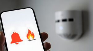 Smartphone displaying fire and bell icons with a smoke detector in the background, symbolizing smart home safety and fire alarm notifications.